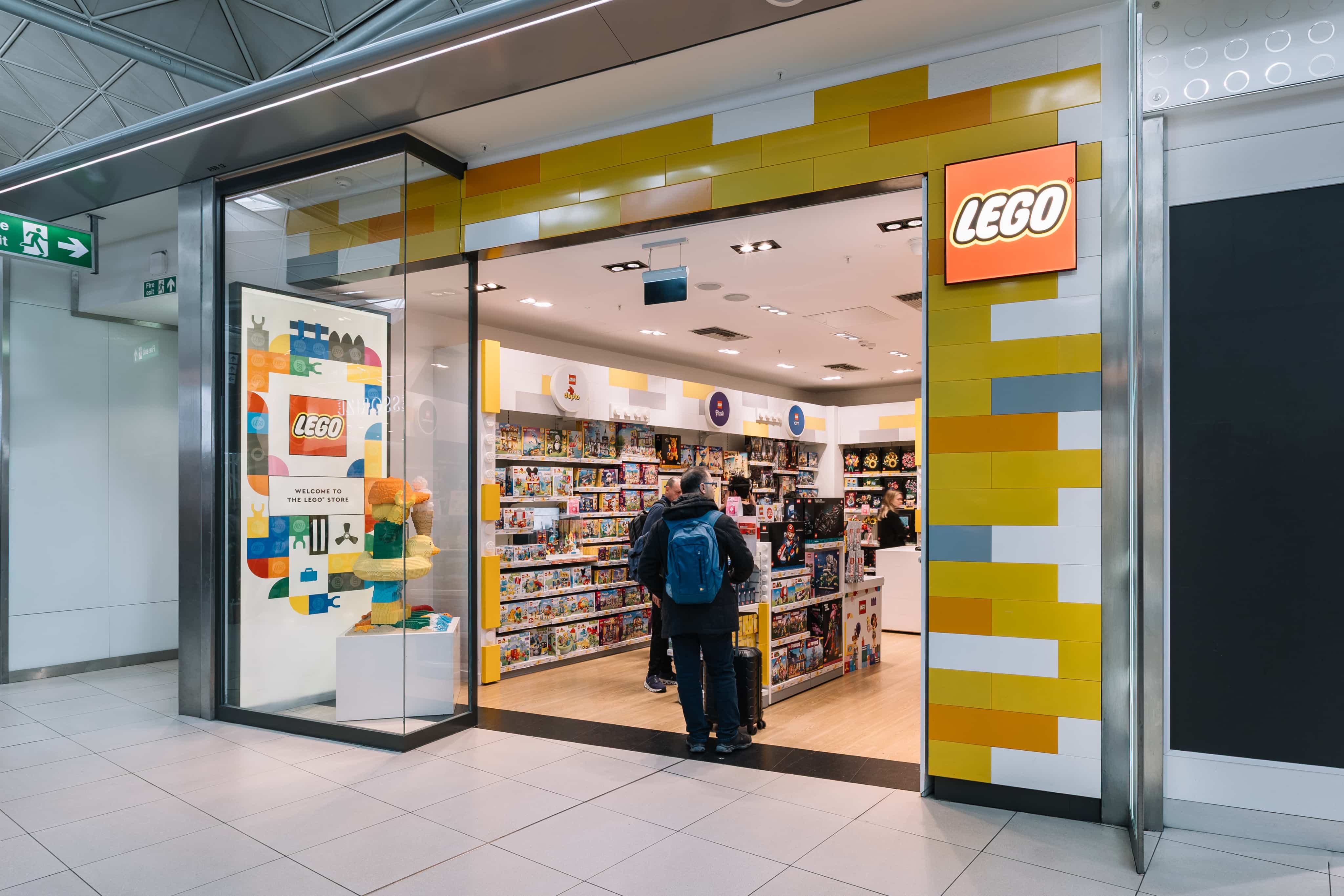 LEGO store entrance with colourful brick-themed décor, shelves full of LEGO sets, and people browsing inside.