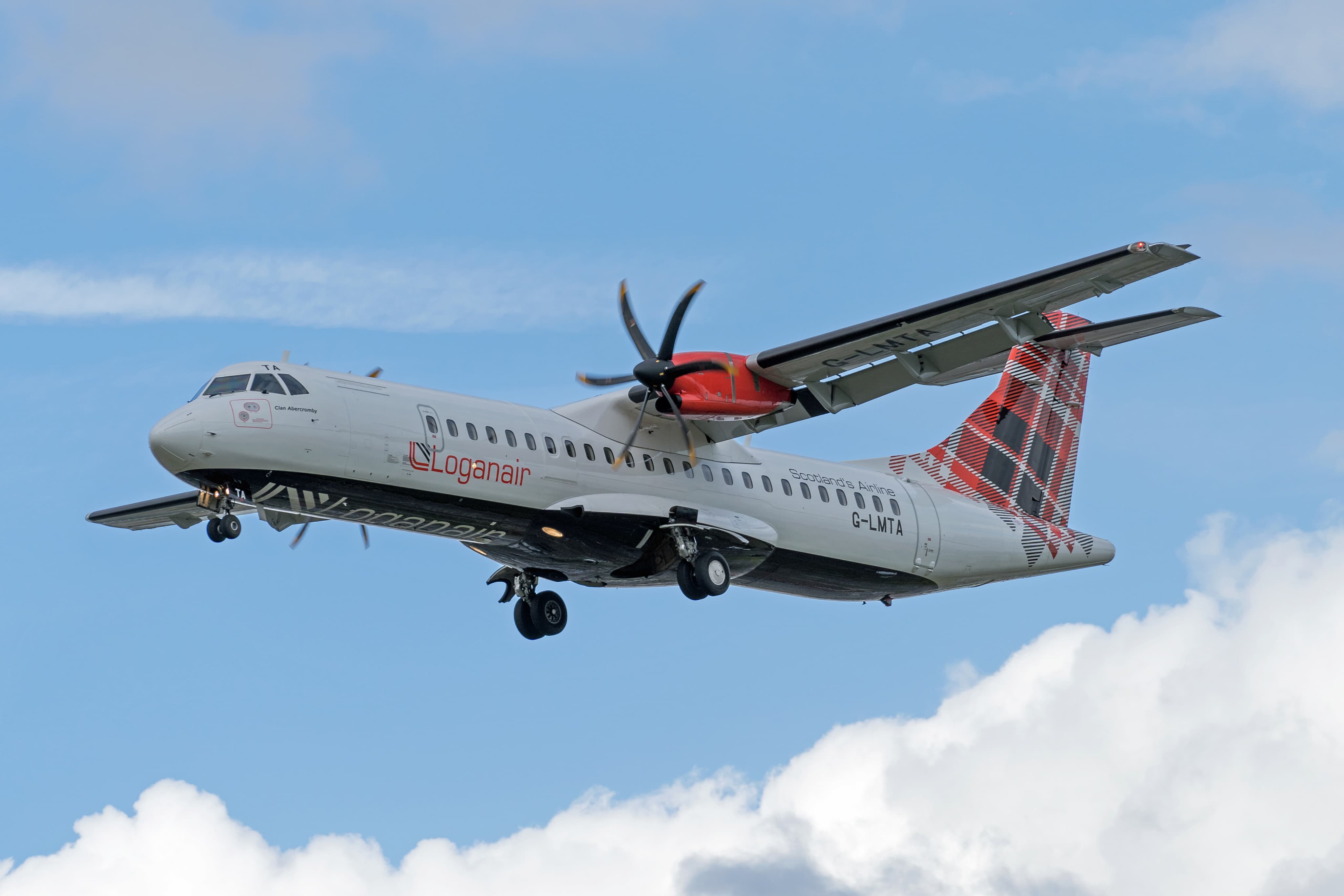 A Loganair ATR aircraft with spinning propellers flying against a blue sky with some clouds.