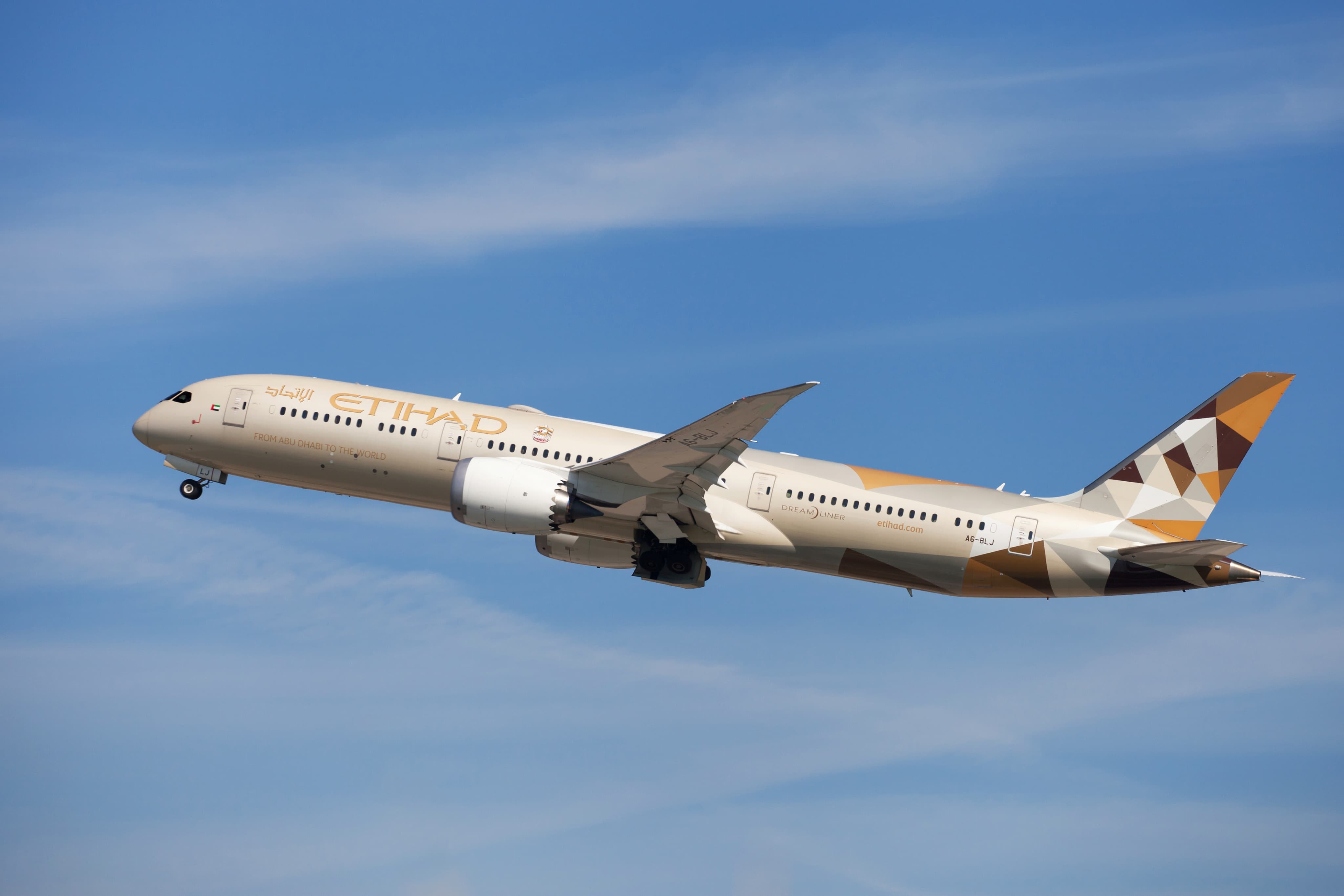 A beige and gold Etihad Airways airplane in flight against a clear blue sky.