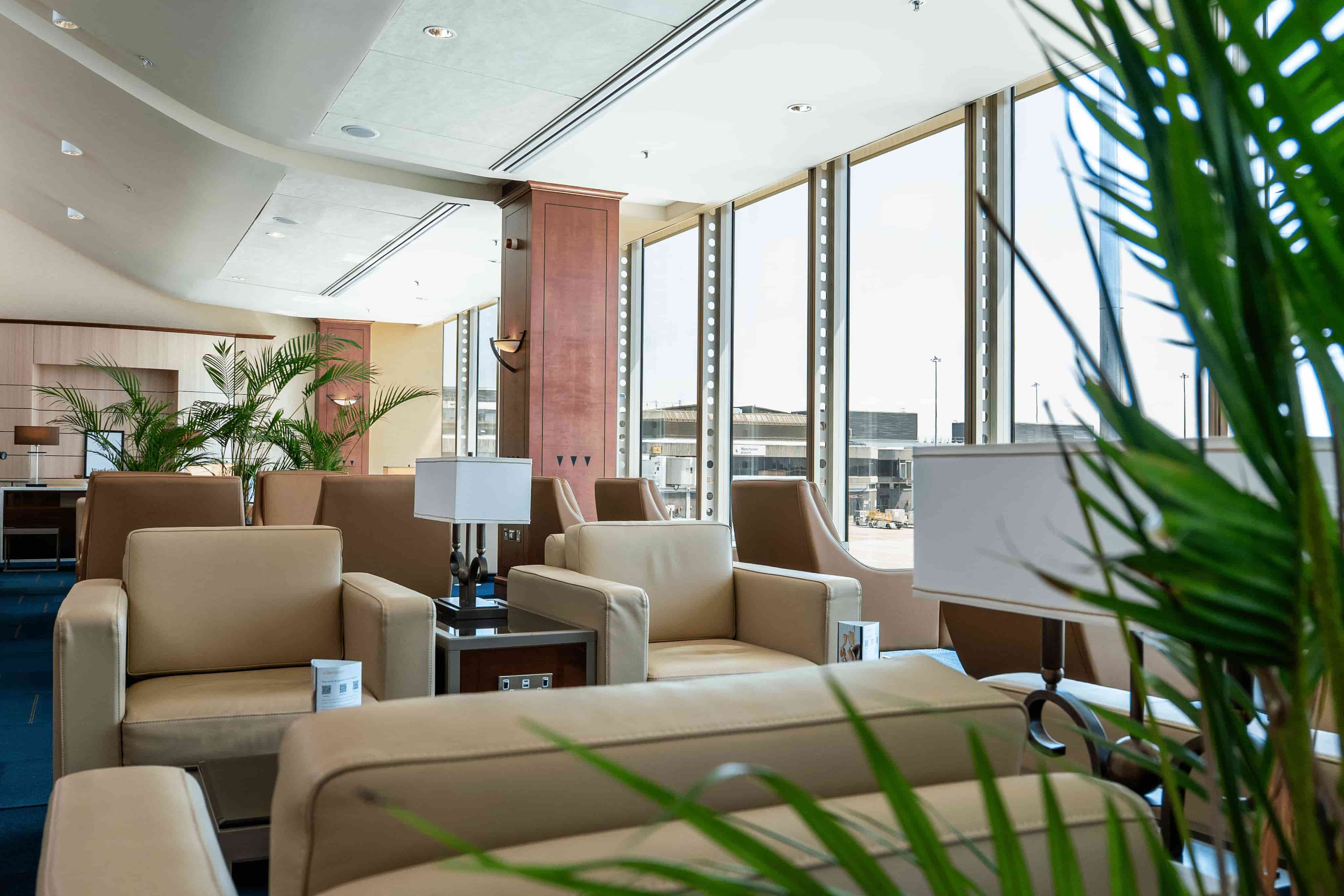 Modern airport lounge with beige leather seating, large windows overlooking the tarmac, and green plants adding a touch of nature.