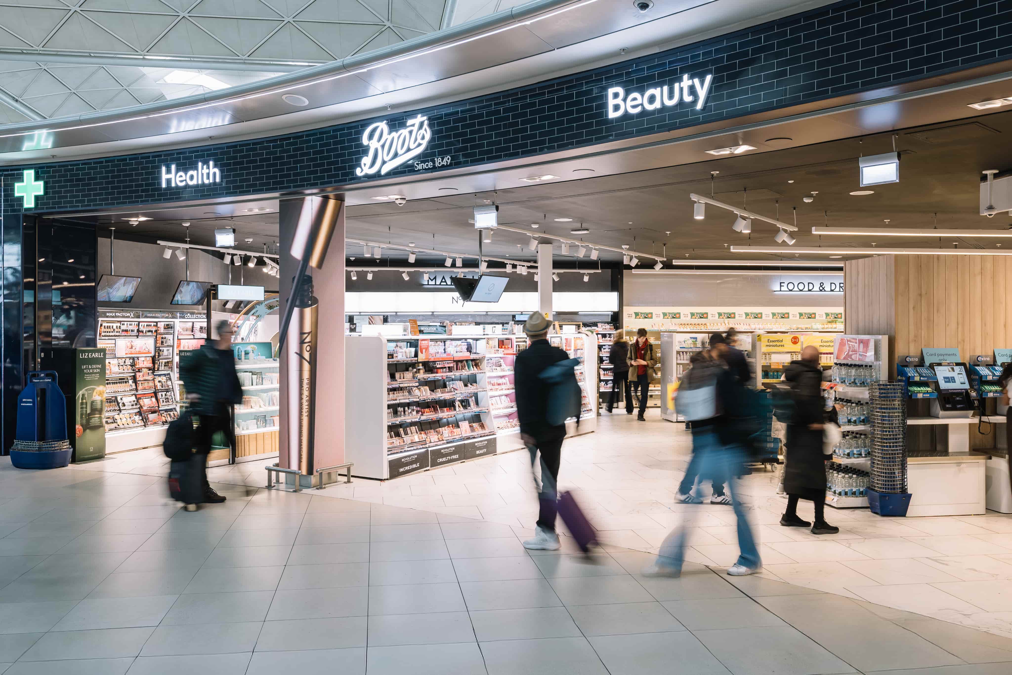 People walking into a brightly lit Boots store located in a spacious area, with signs for health and beauty products.