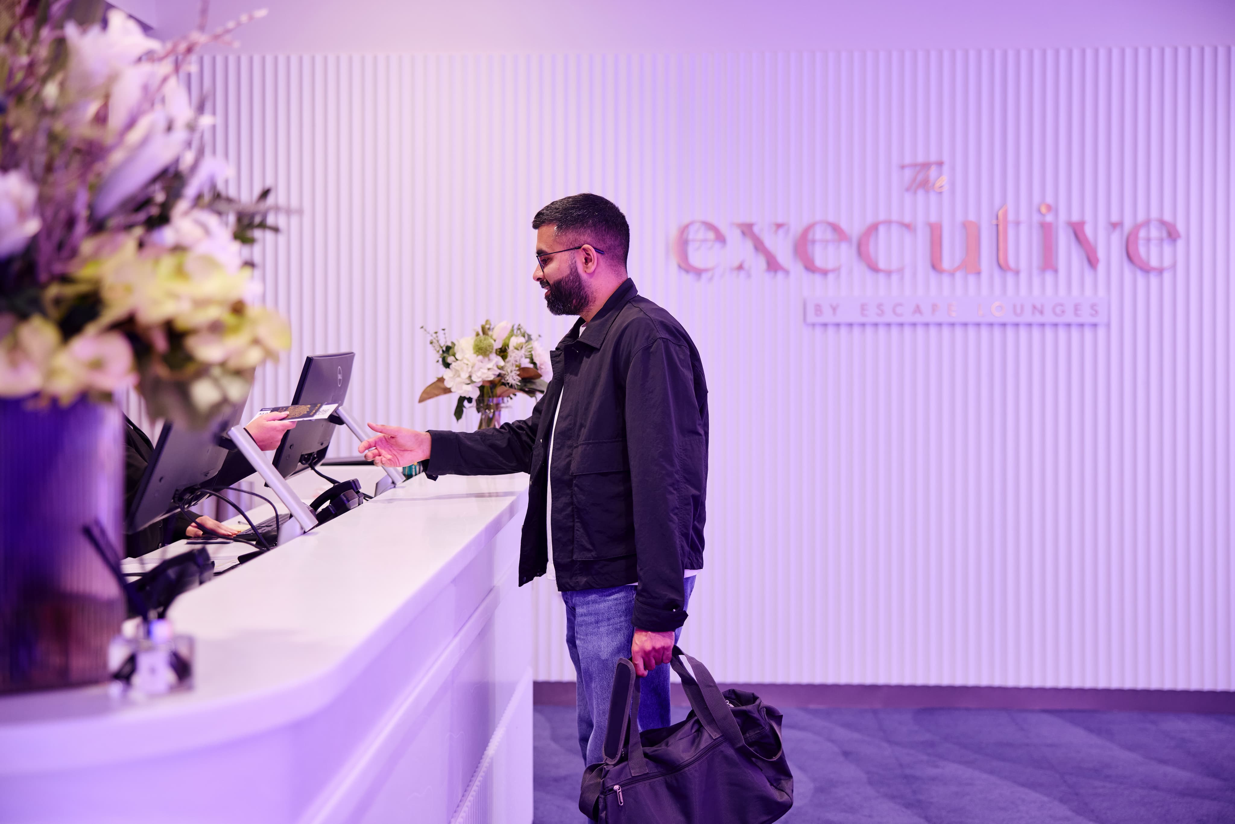 A man at a hotel reception counter checks in, handing over an ID while holding a bag. The background shows the logo "The Executive."