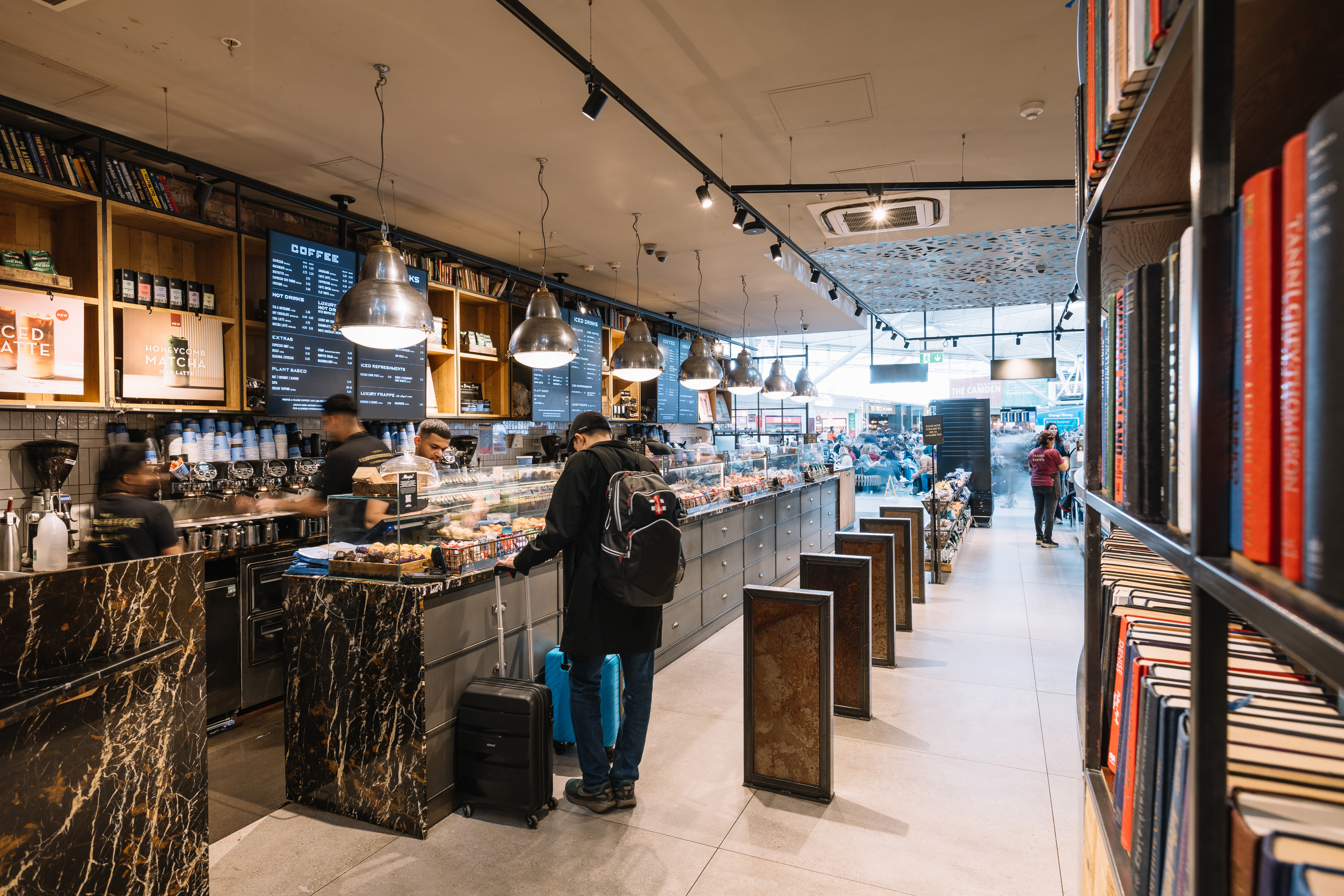 A cosy coffee shop with a marble counter, customers ordering drinks, hanging lights, and shelves filled with books and travel essentials.