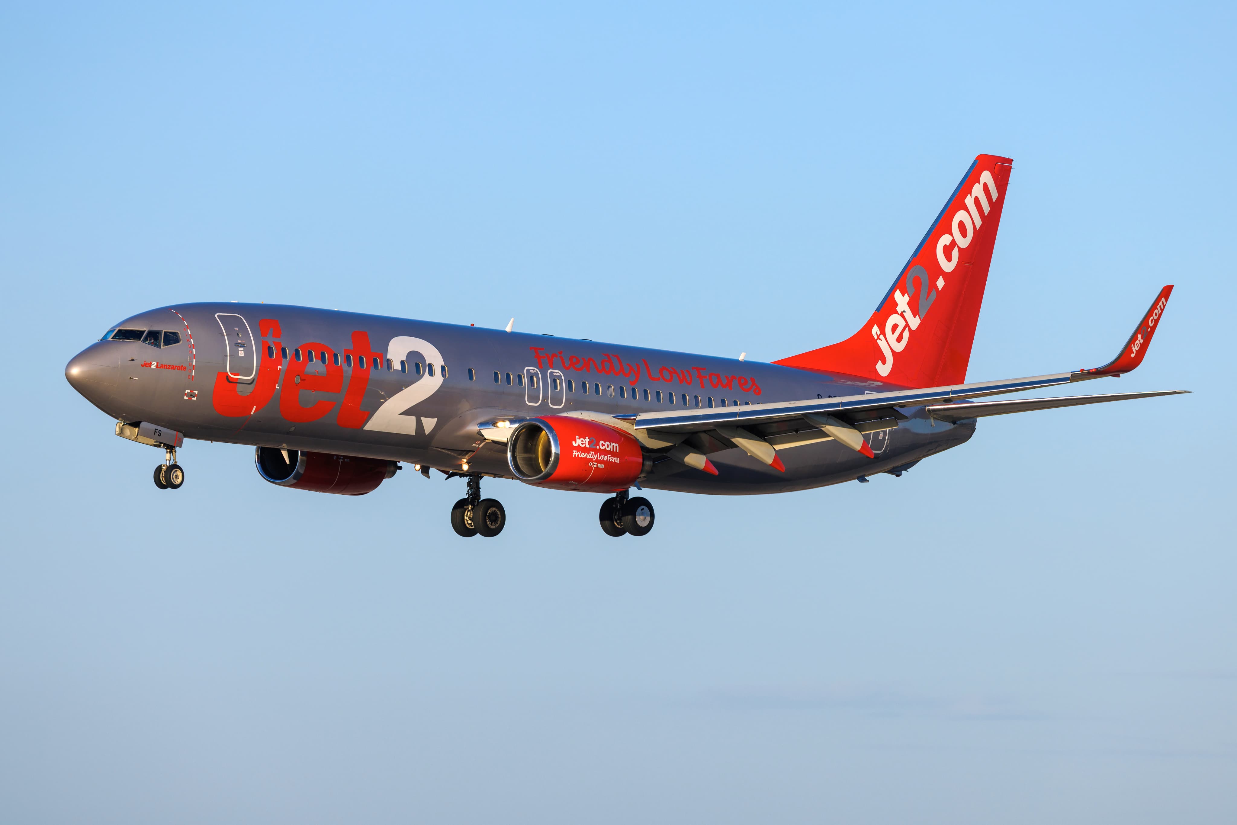 A Jet2 airplane in flight, displaying its silver and red livery with the company logo and "Friendly Low Fares" slogan against a clear blue sky.