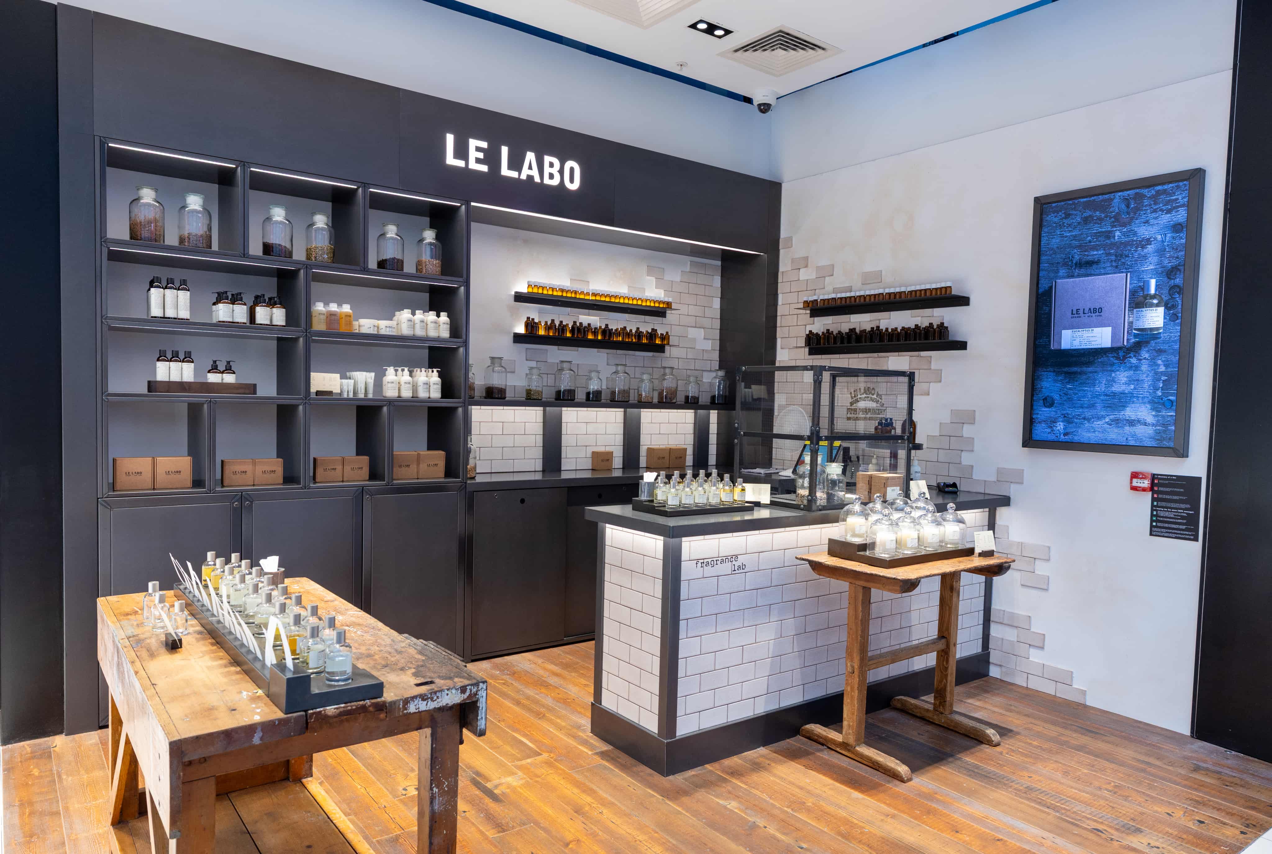 A perfume store interior featuring shelves with various bottles and products, a wooden display table, and a modern design with tiled walls.