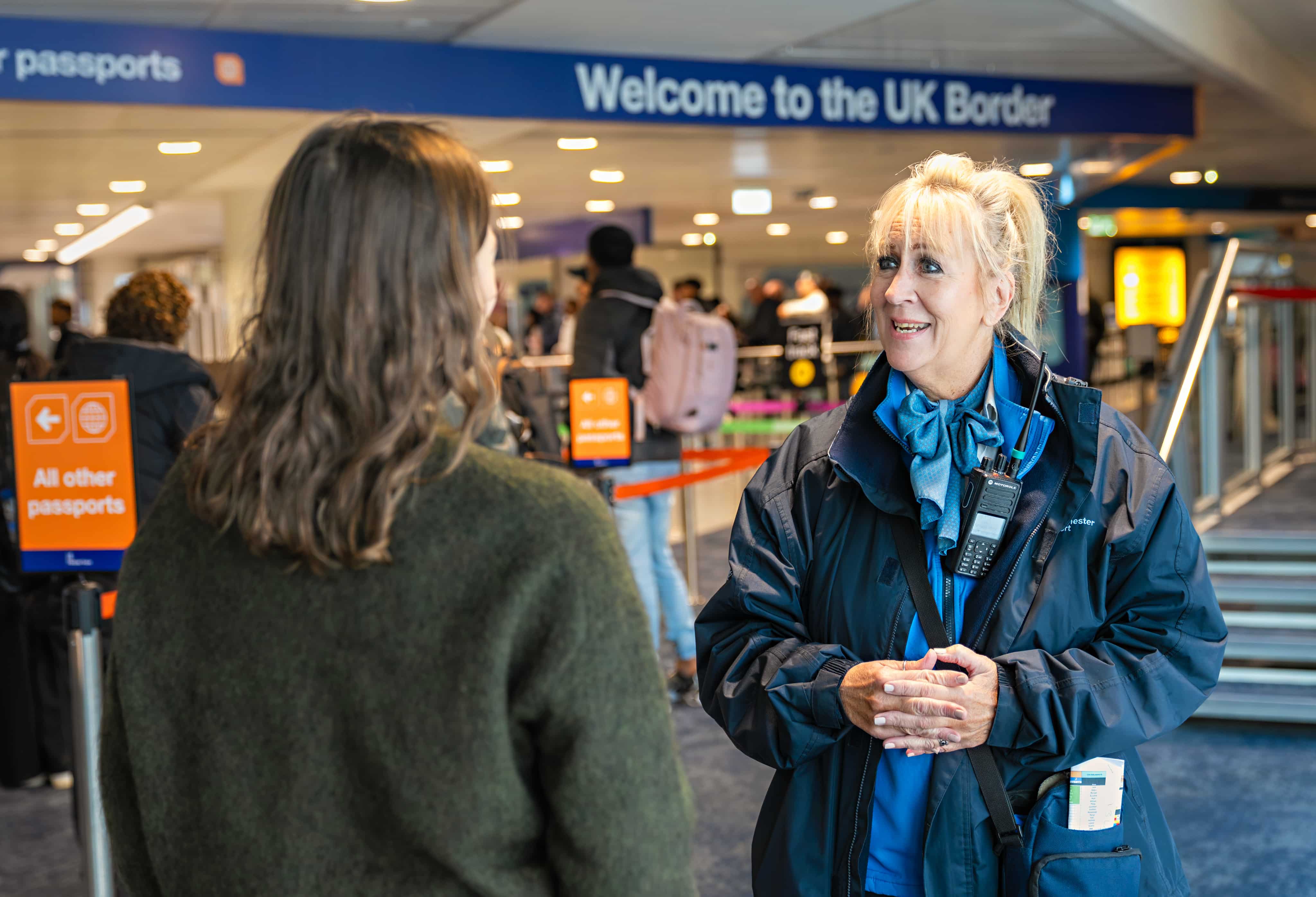 A security staff member, in uniform, smiles while speaking to a traveller at the UK Border, with signs for passport queues visible.