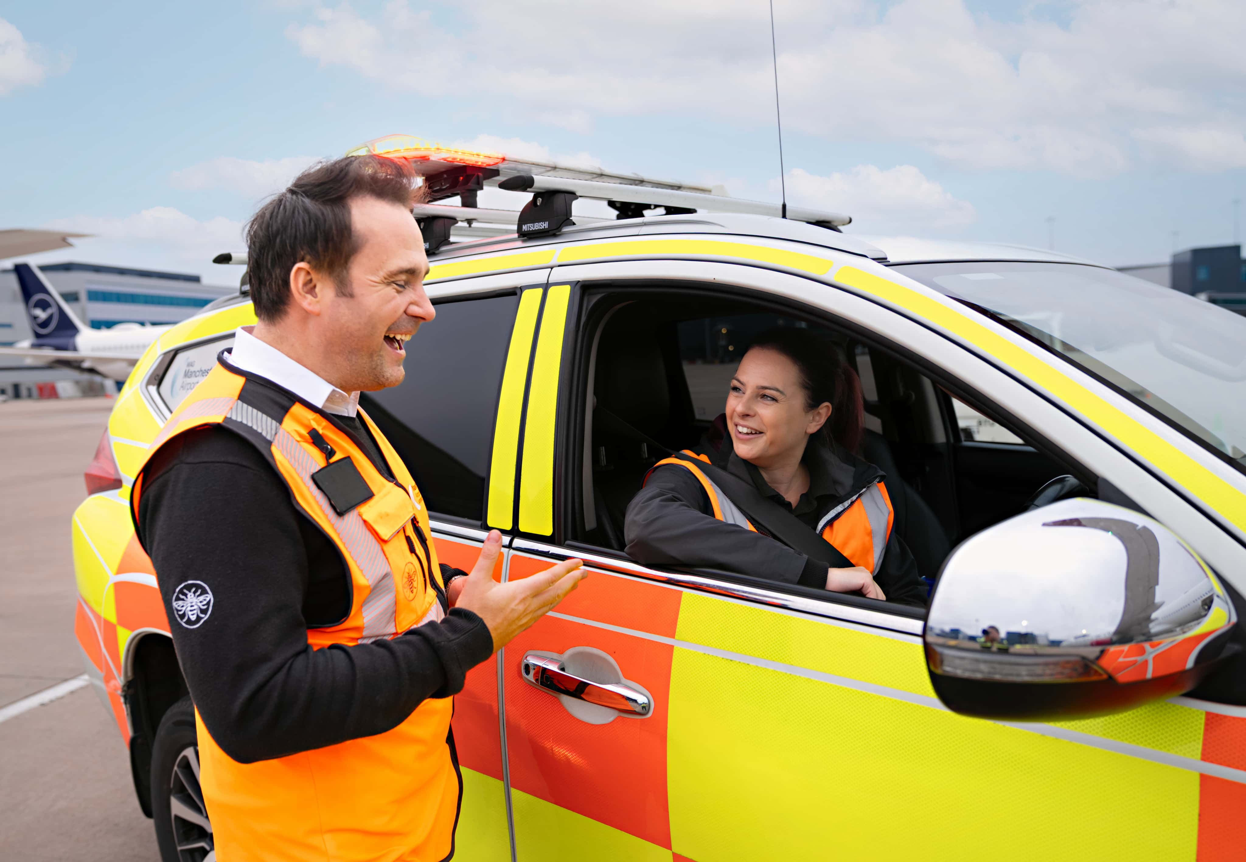 Two airport personnel in orange safety vests chat by a yellow vehicle on the tarmac, with a plane and terminal in the background.