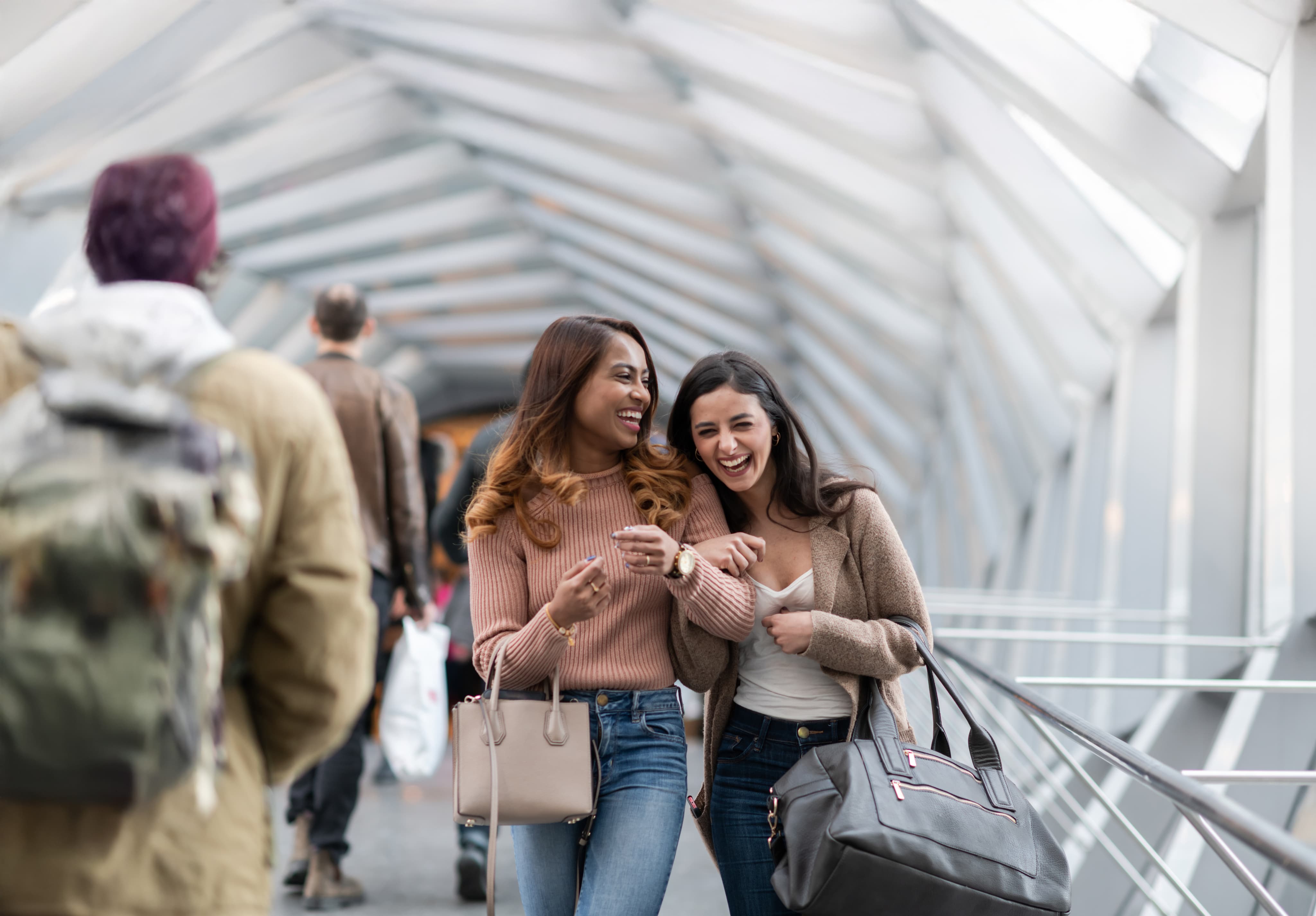 Two women walking and laughing in a covered walkway. Both are casually dressed, carrying handbags, with people in the background.