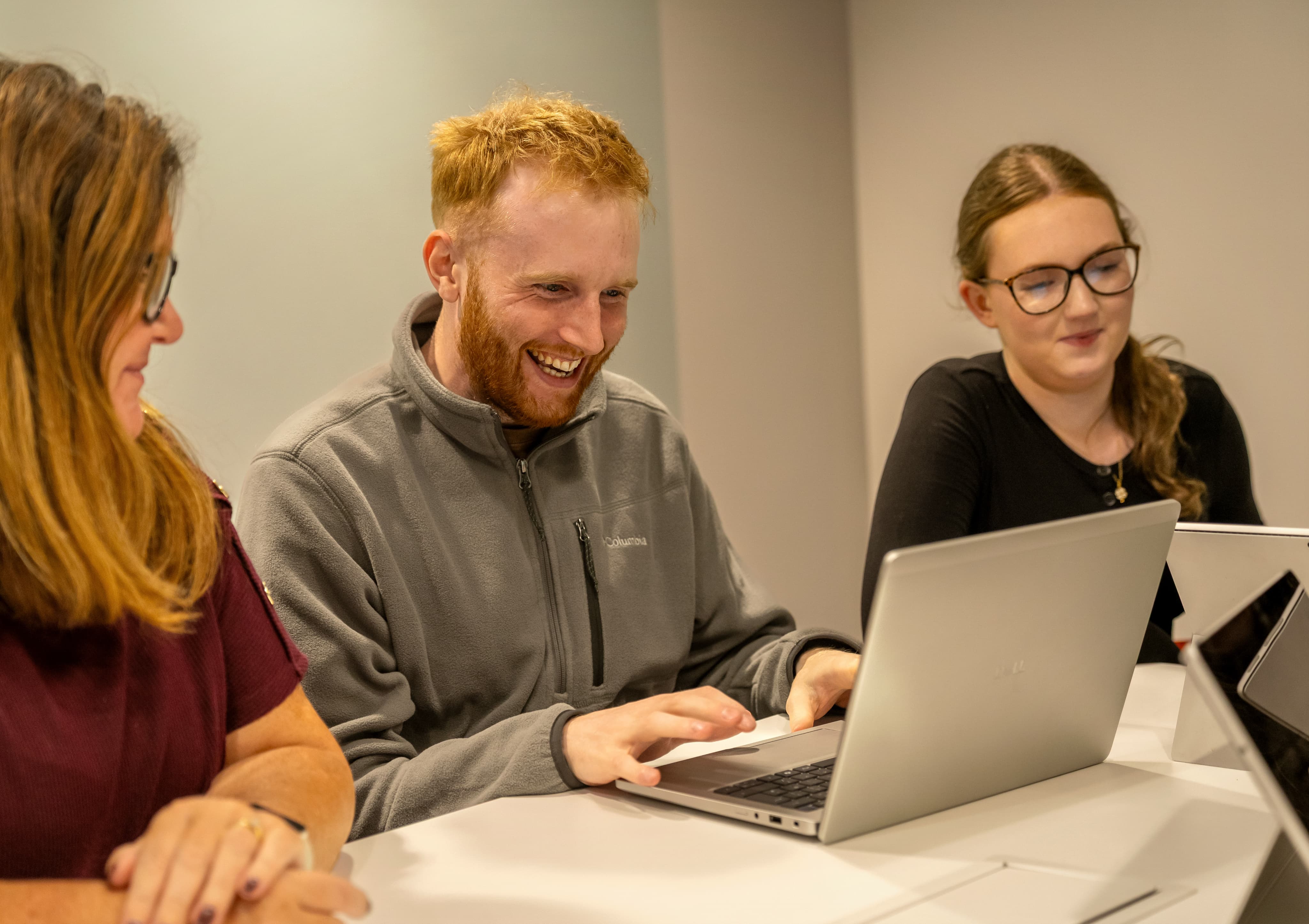 Three people smiling and working on laptops at a table, appearing engaged in a friendly discussion.