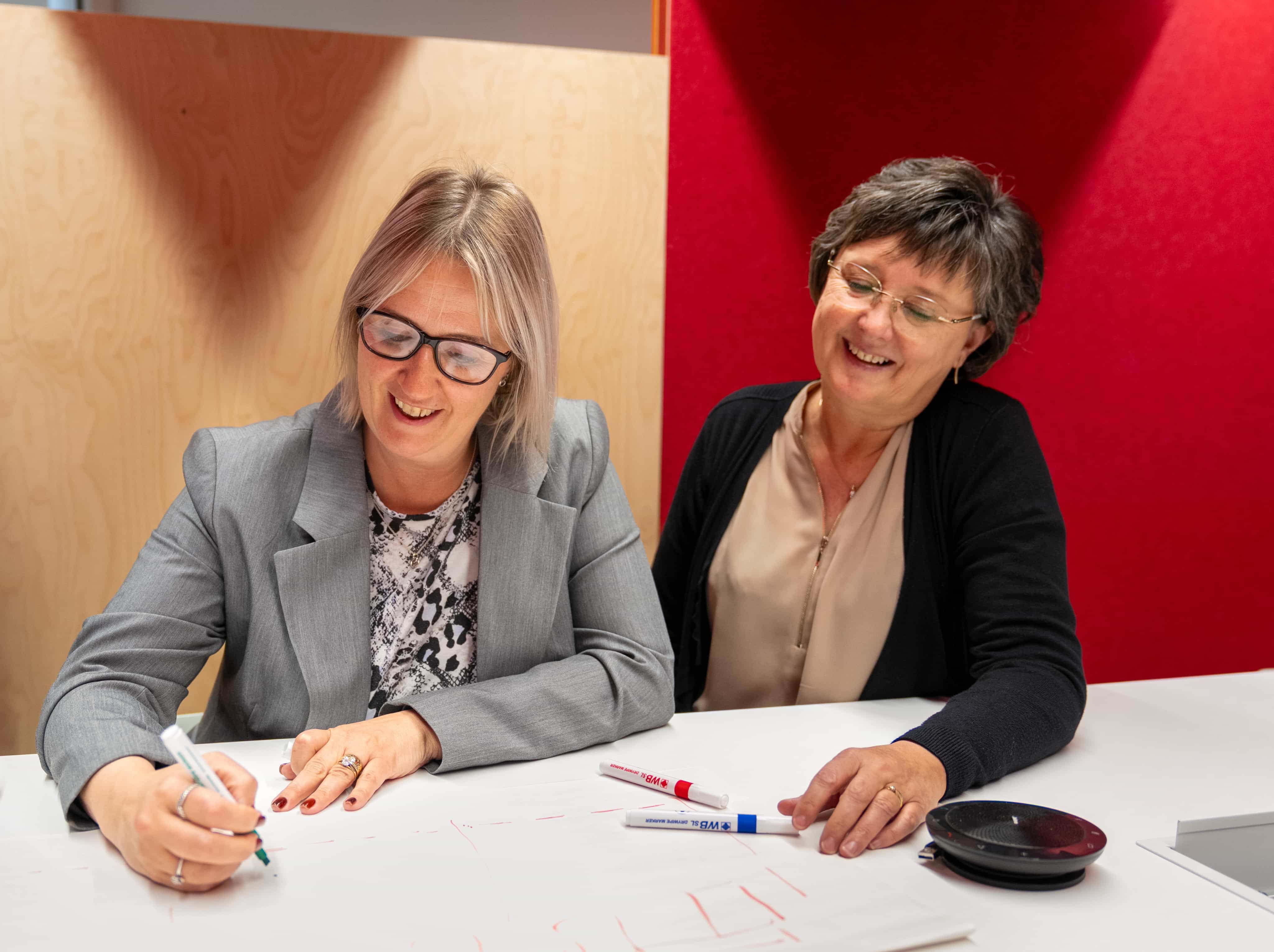 Two women smiling and collaborating at a table, one writing with a marker. Papers and additional markers are visible.