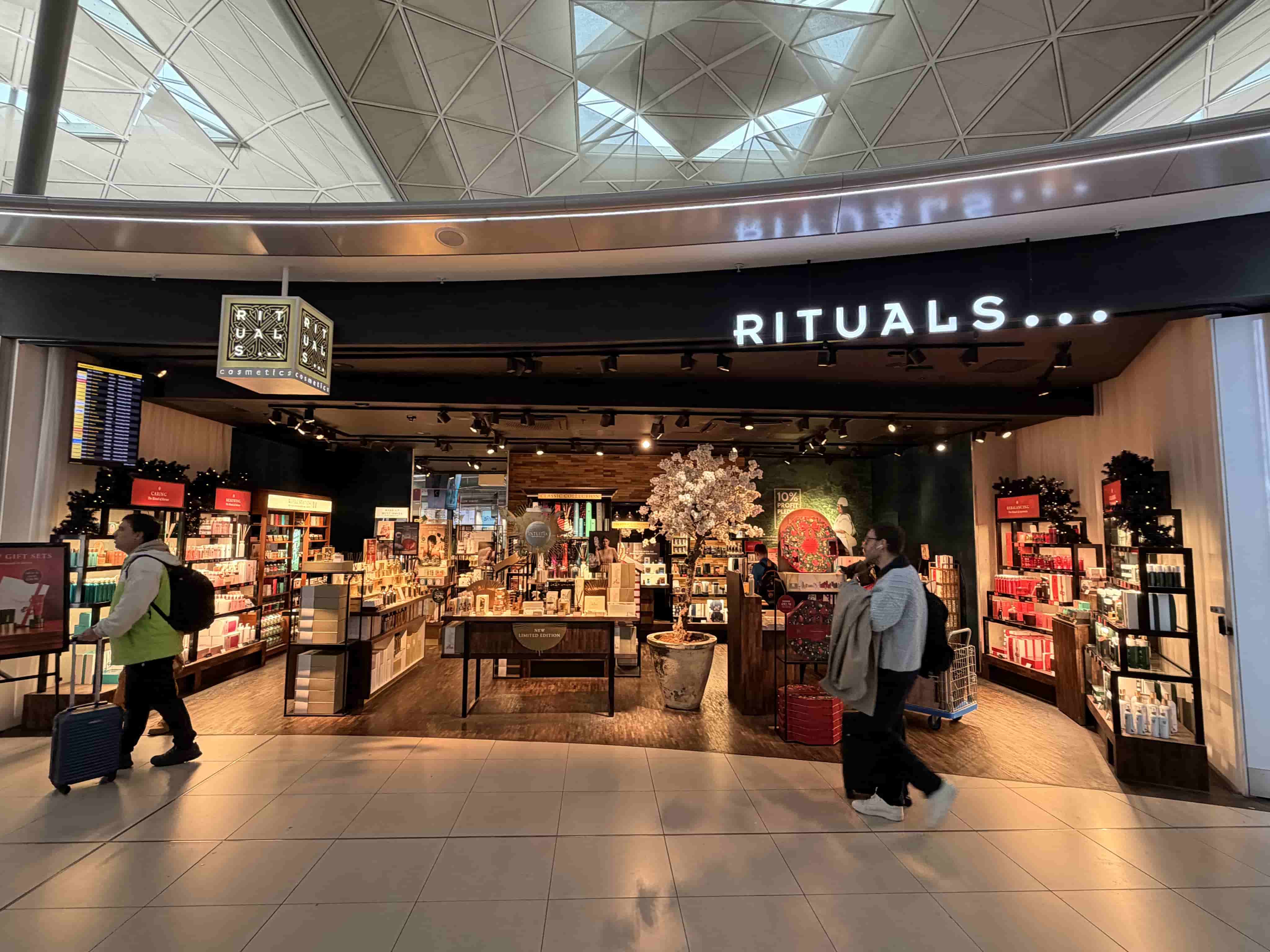 Rituals cosmetics store entrance in an airport concourse, illuminated sign, product displays and travelers passing with luggage.