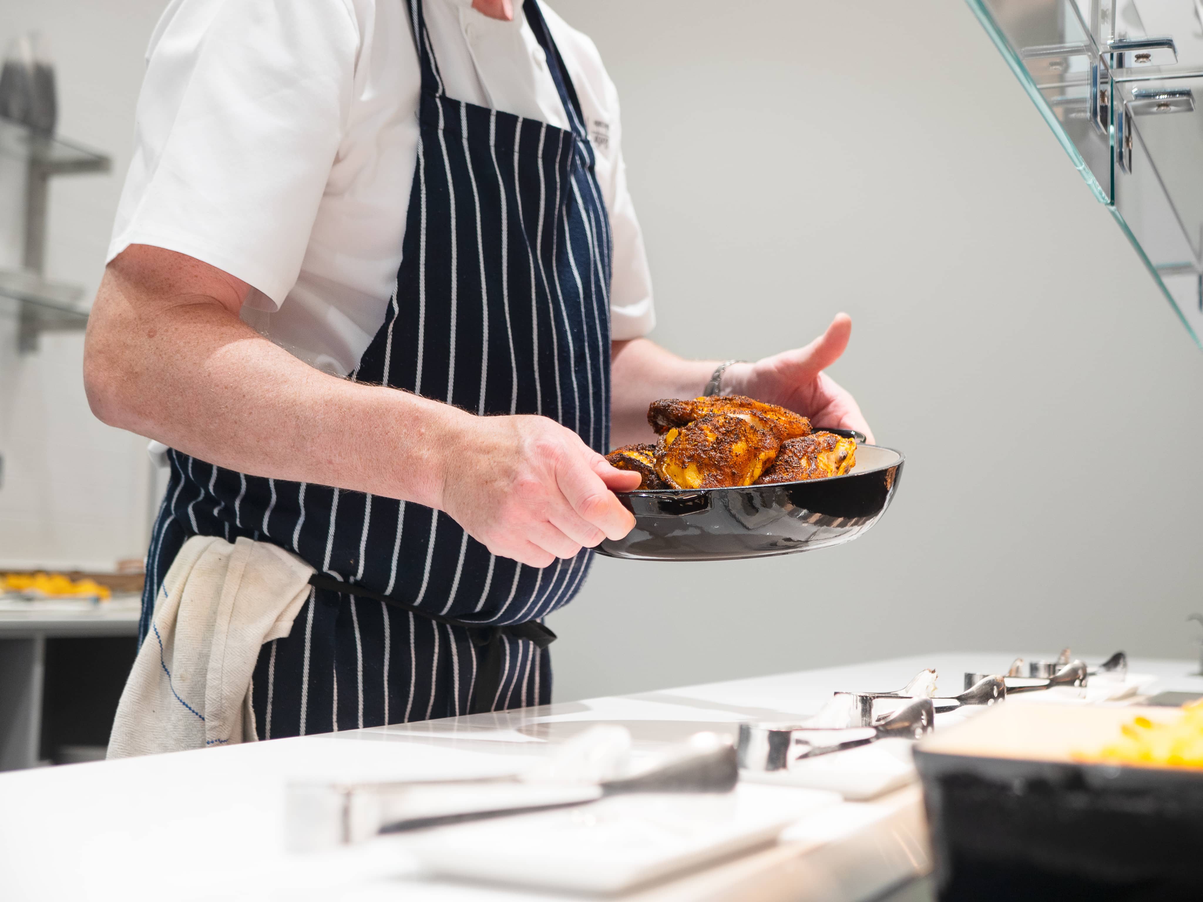 Chef in a striped apron placing a cooked chicken on a counter in a restaurant kitchen.
