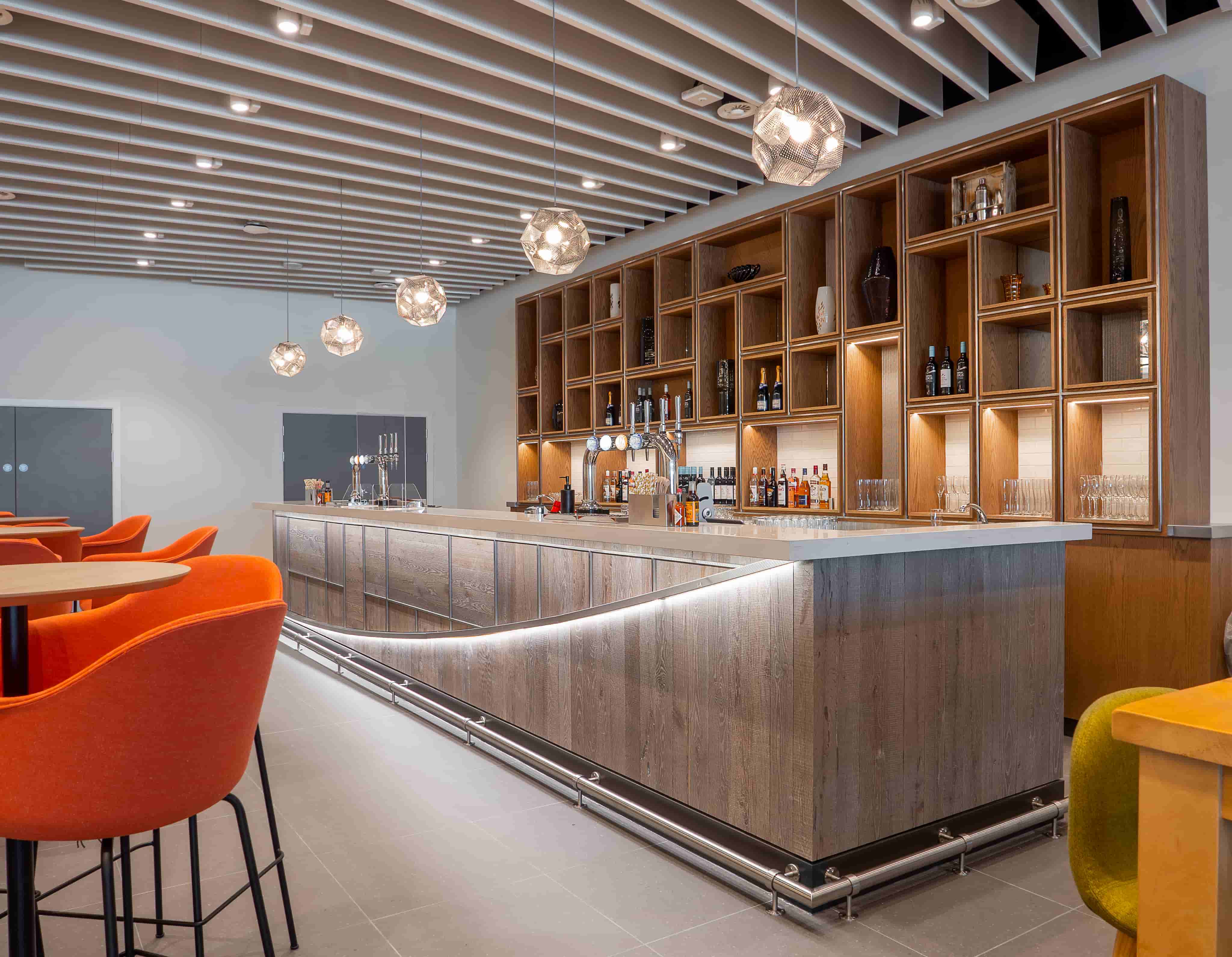 Modern bar with a wooden counter, orange chairs, pendant lights, and shelves filled with bottles and glassware.