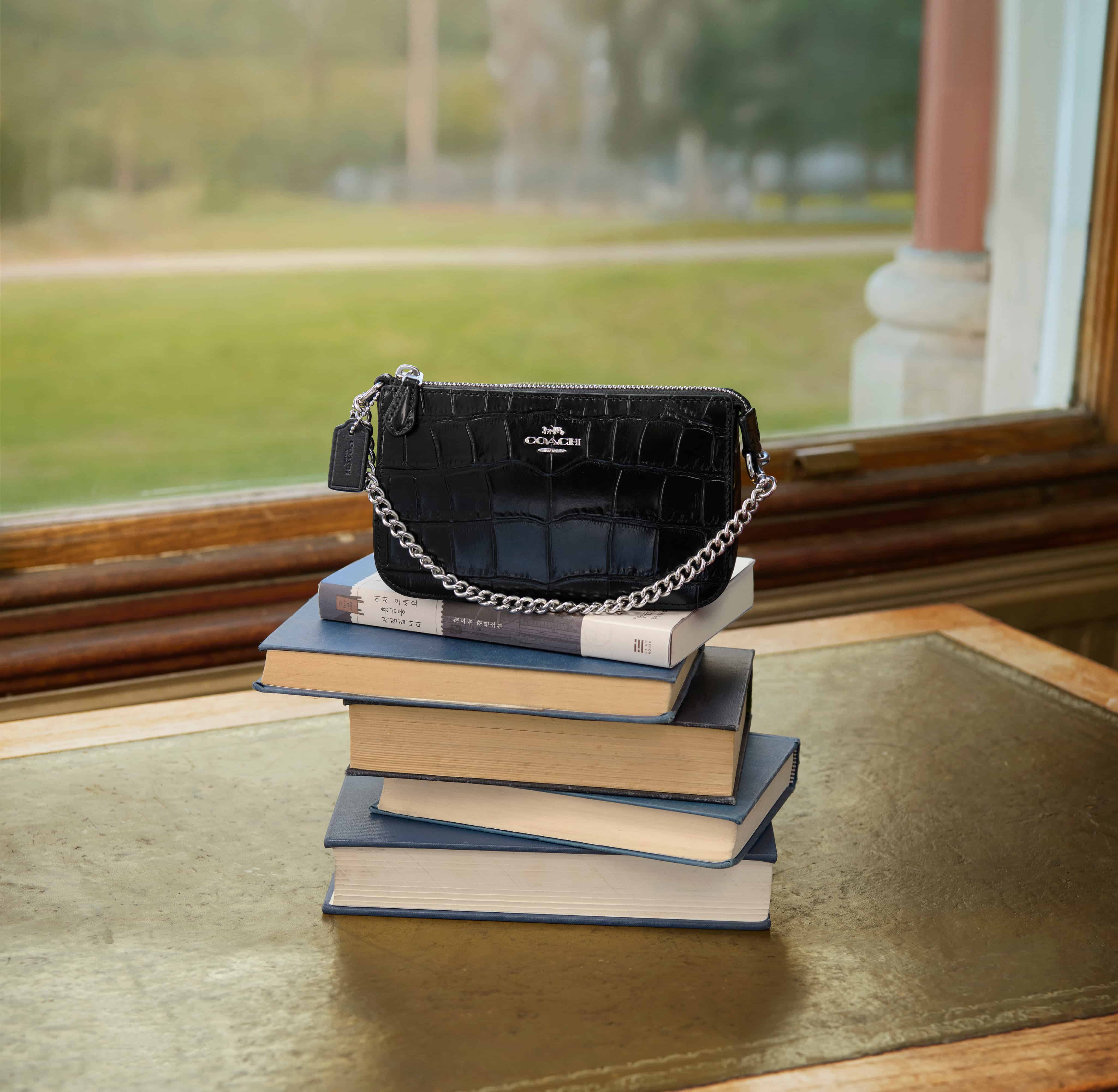A black textured handbag with a chain strap rests on a stack of books on a table, with a window view of a grassy field in the background.