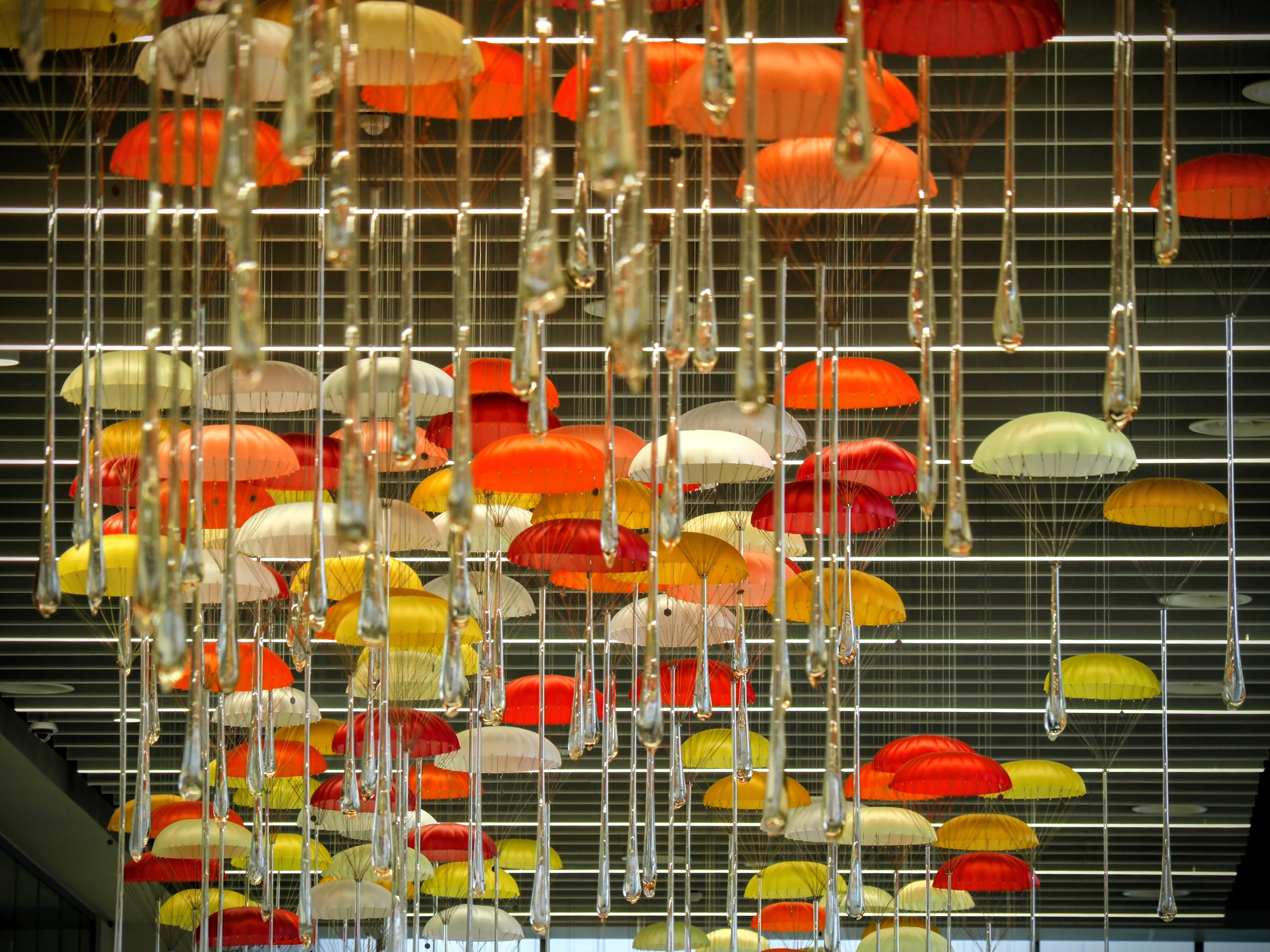 Colourful umbrellas and teardrop-shaped glass hang from the ceiling, creating a vibrant art installation against a grid-patterned background.