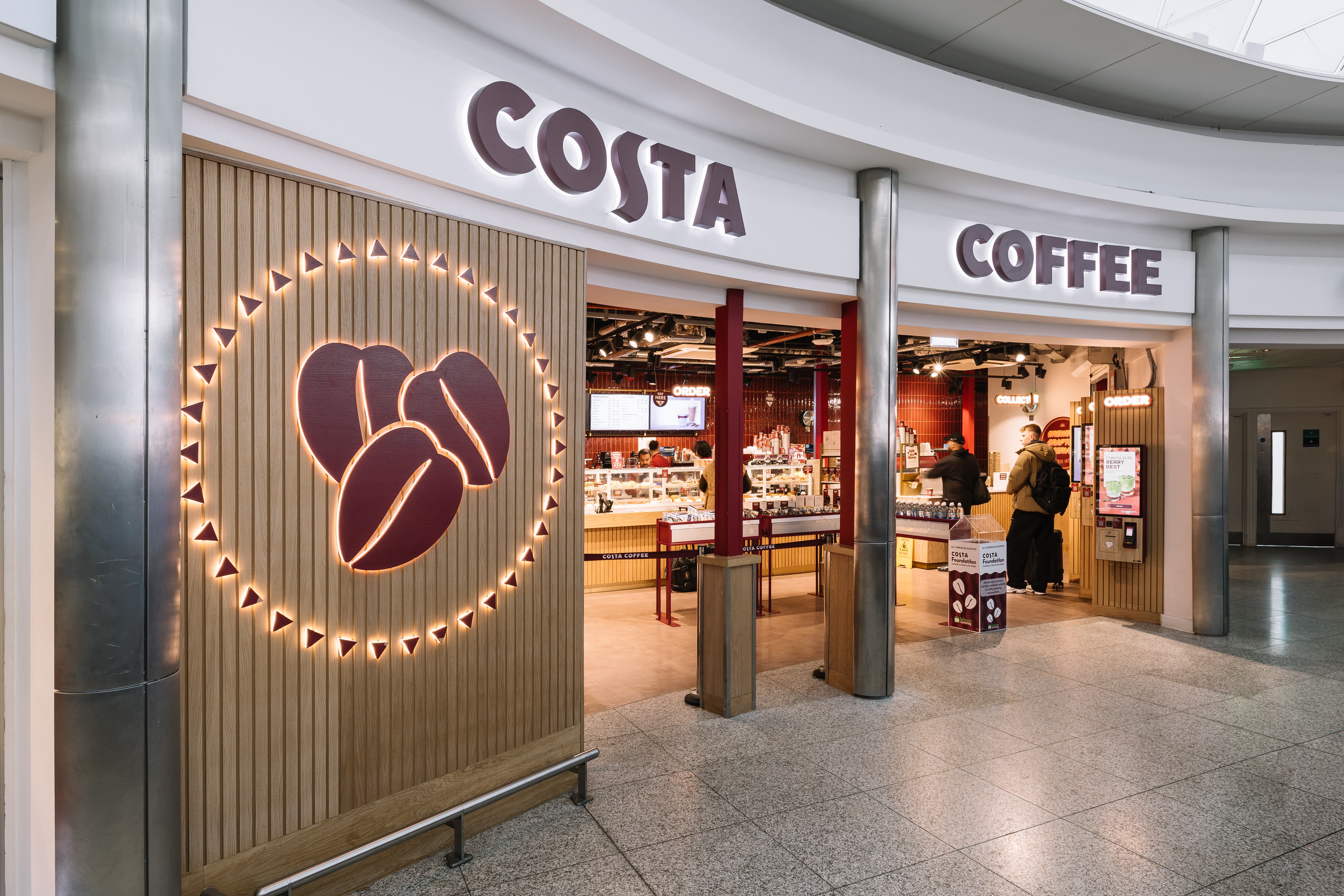 Costa Coffee shop entrance with illuminated coffee bean logo, modern décor, and customers ordering at the counter inside.