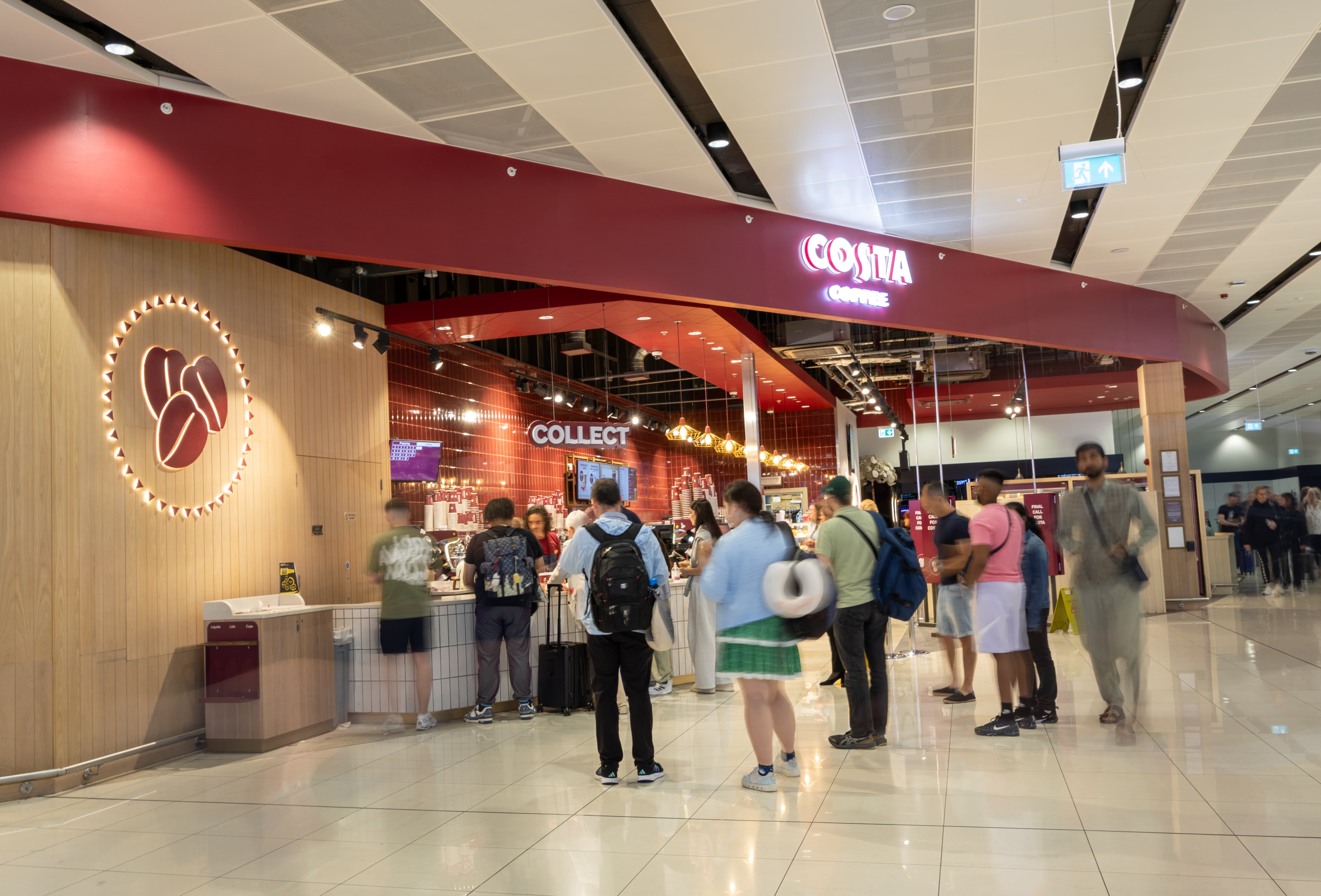 People wait in line at a Costa Coffee shop in a modern, brightly lit setting with a red and wood decor theme