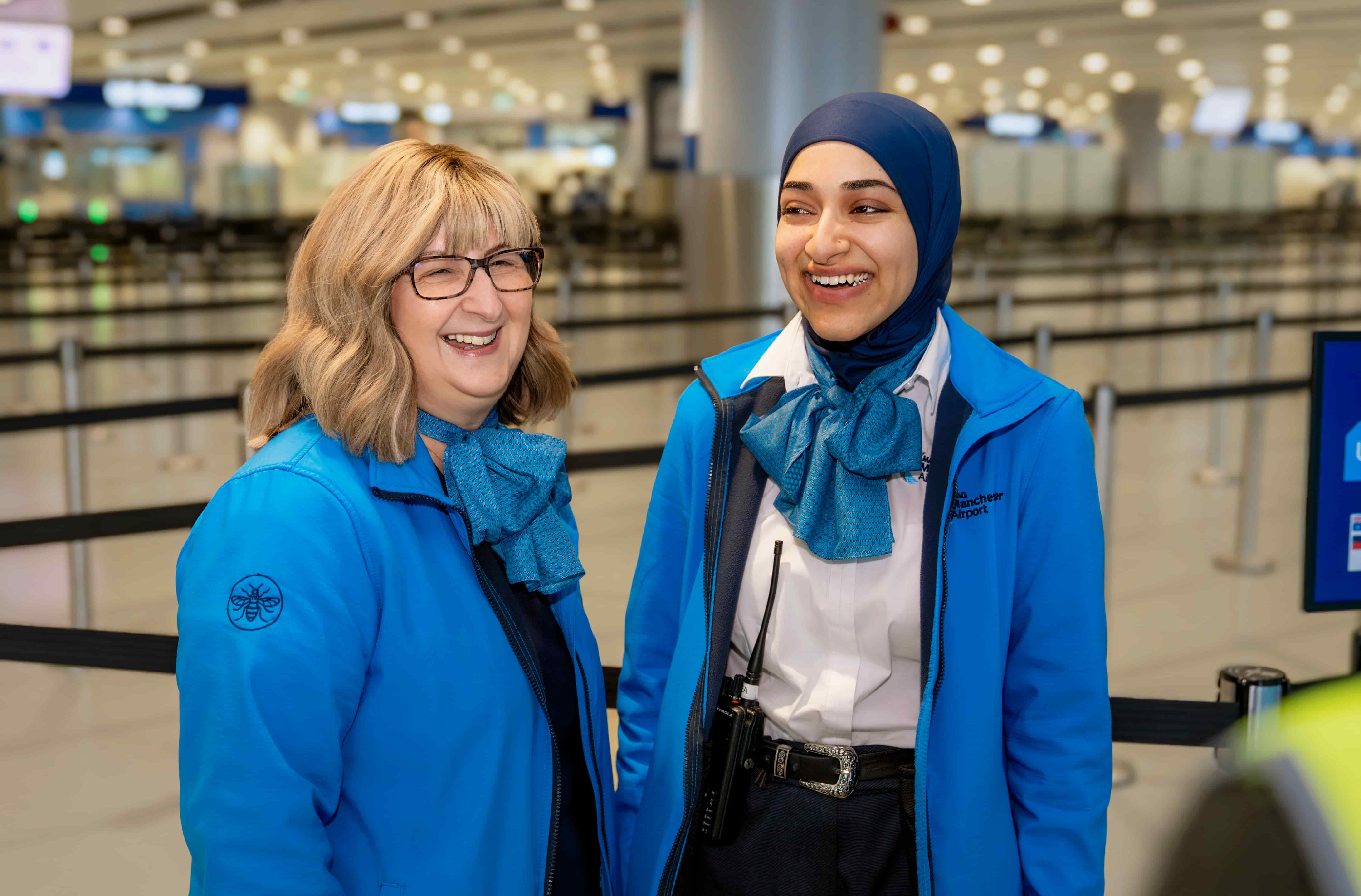 Two smiling airport staff in blue uniforms and scarves stand inside a terminal, with check-in counters and lane dividers in the background.