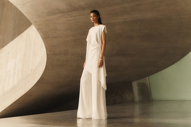 A woman in a sleek white, sleeveless dress poses in a modern, minimalist architectural setting with curved, concrete walls.