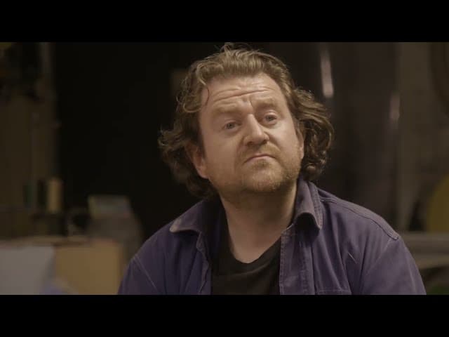 Liam Hopkins, an artist with curly hair and a beard wearing a blue shirt and black undershirt, looking slightly to the side, in a dimly lit room.