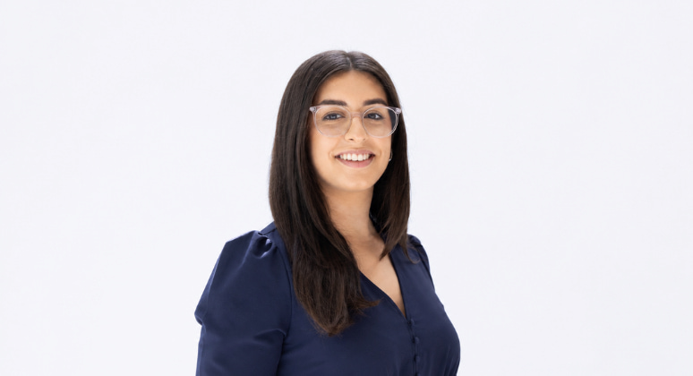 A woman with long dark hair and glasses smiles, wearing a navy blouse against a white background.