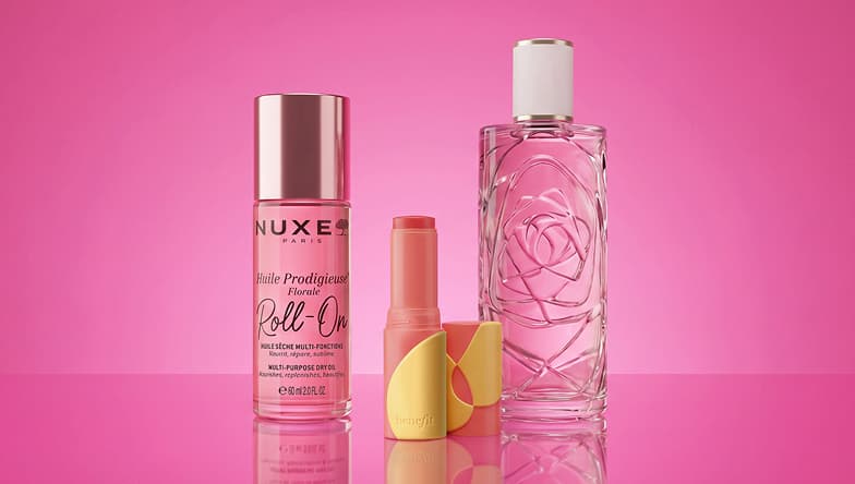 Three cosmetic products on a pink background: a Nuxe roll-on, a pink lipstick with yellow cap, and a perfume bottle with a rose design.