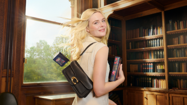 Woman with long blonde hair smiling, holding a book in a library with bookshelves, wearing a beige dress and black purse. Sunlight streams through the window.