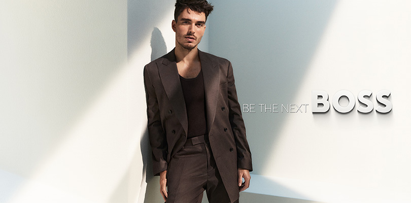Man in a sleek suit stands in sunlight against a white wall with "Be the Next Boss" text beside him.