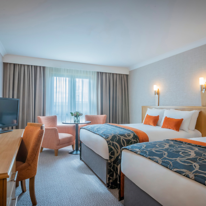 Modern hotel room with two twin beds, orange pillows, stylish chairs, wall sconces, a small table, and a TV on a wooden desk.