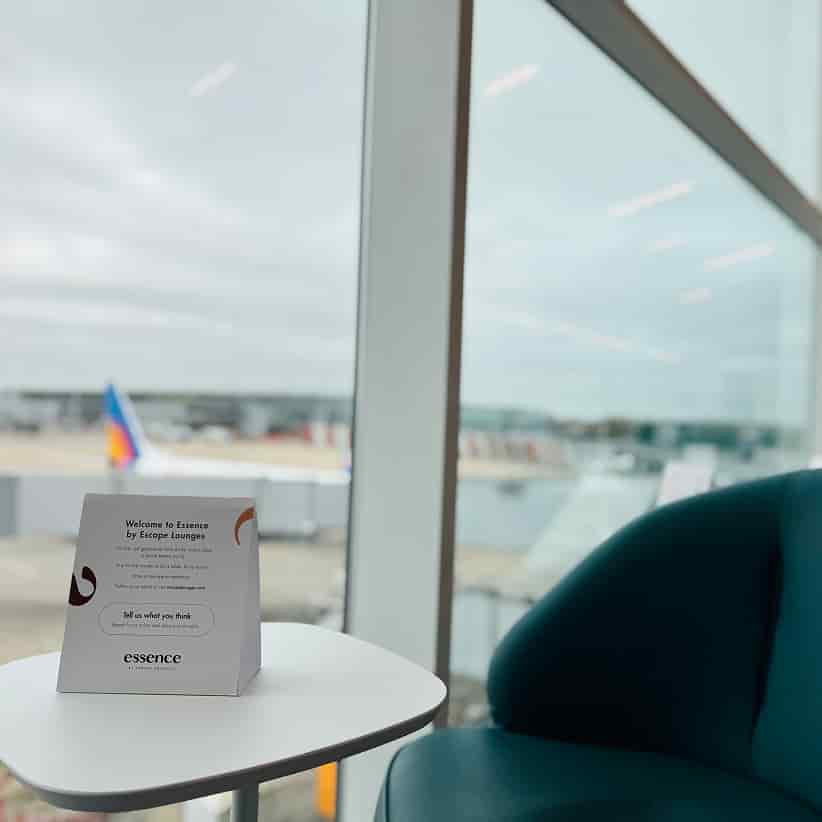 A lounge area with a sign reading "Welcome to Essence by Escape Lounges" on a small table, and an airport runway visible through the window.