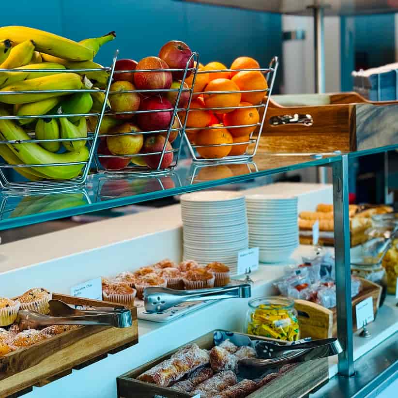 A buffet with baskets of bananas, apples, oranges, and trays of pastries. Plates and utensils are neatly arranged, with snacks and muffins displayed.