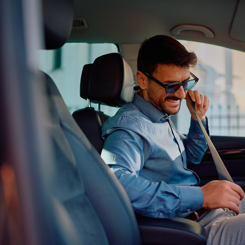 A man in sunglasses and a blue shirt fastens his seatbelt while seated in a car.